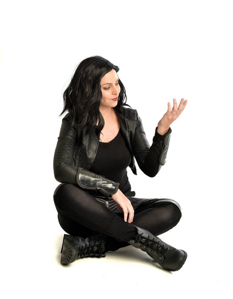 Ganzkörperporträt eines schwarzhaarigen Mädchens im Lederkostüm. sitzend in Pose, isoliert auf weißem Studiohintergrund. - Foto, Bild