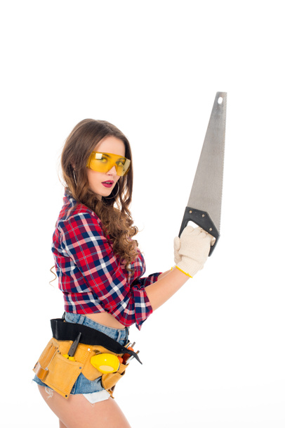 beautiful sexy girl in goggles with tool belt holding saw, isolated on white - Fotó, kép