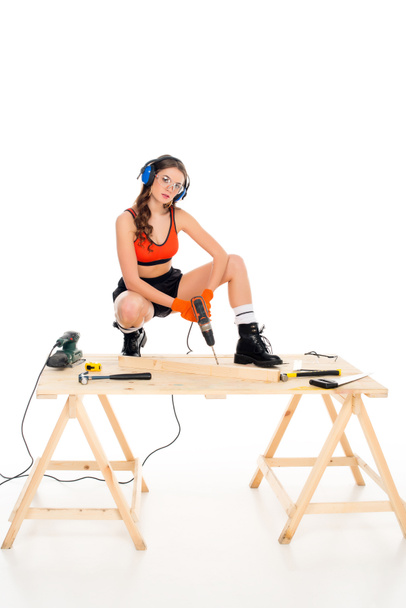 girl in protective headphones working with electric drill at wooden table with tools, isolated on white - Photo, Image