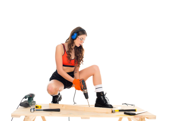 сексуальная девушка в защитных наушниках, работающая с электрической дрелью за деревянным столом с инструментами, изолированными на белом
 - Фото, изображение