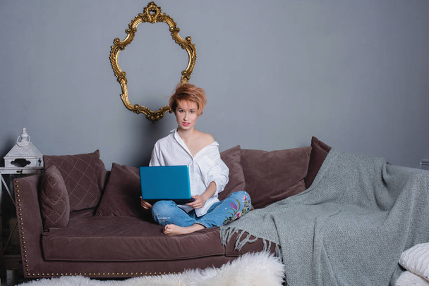 Stylish young woman with laptop in hands on sofa and looking at camera . Dressed in a white shirt and jeans with embroidery. Gray wall with shadow behind her. - Photo, image