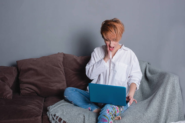 Stylish surprised young woman with laptop in hands on sofa and looking at camera . Dressed in a white shirt and jeans with embroidery. Gray wall with shadow behind her. - Foto, Imagen