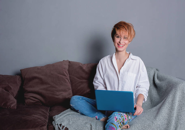 Stylish young woman with laptop in hands on sofa and looking at camera . Dressed in a white shirt and jeans with embroidery. Gray wall with shadow behind her. - Foto, Bild