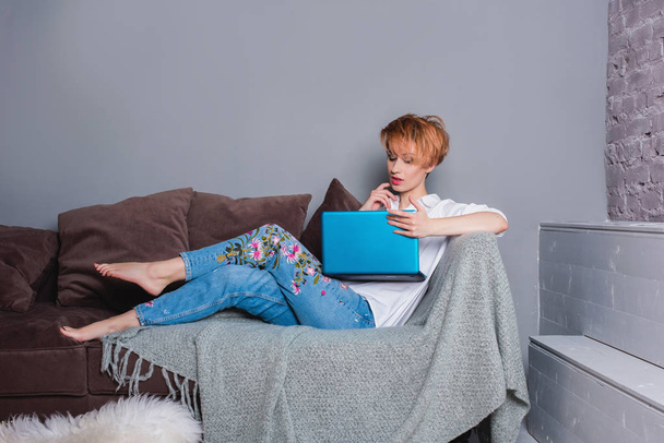 Stylish young woman with laptop in hands on sofa and looking at laptop . Dressed in a white shirt and jeans with embroidery. Gray wall with shadow behind her. - Zdjęcie, obraz
