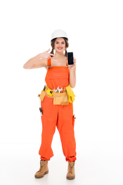 smiling workwoman in uniform pointing at smartphone with blank screen, isolated on white - Zdjęcie, obraz