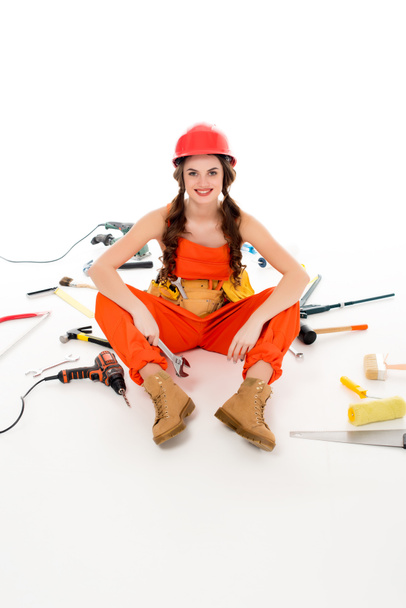 smiling girl in overalls and hardhat sitting on floor with different tools, isolated on white - Foto, Imagen