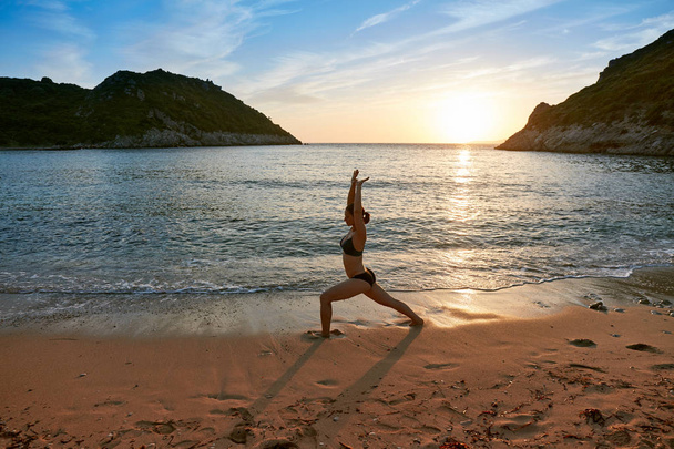Sylwetka praktyki jogi o zachodzie słońca. Młoda kobieta robi ćwiczenia jogi na plaży. Corfu, Grecja. - Zdjęcie, obraz