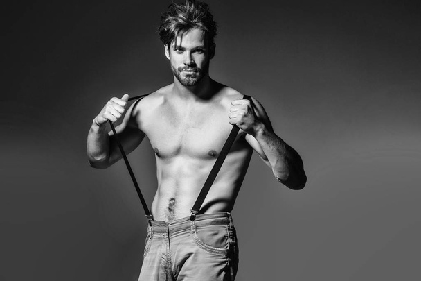 Handsome guy fashion sexy young bearded macho man model with suspenders on pants has bare muscular torso holds on grey background - 写真・画像