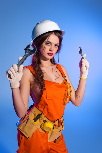 brunette girl in overalls and safety helmet with tool belt holding wrenches, isolated on blue - Foto, Bild