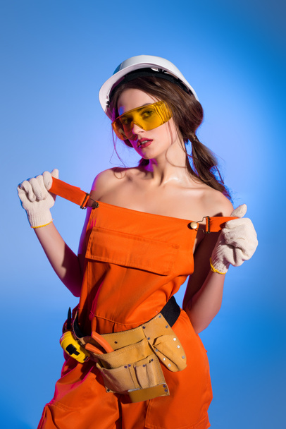 seductive girl in overalls uniform and safety helmet, isolated on blue - Foto, Imagen