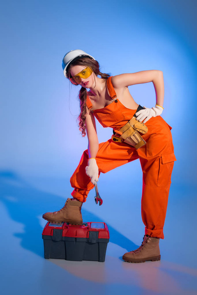 attractive girl in overalls uniform with tool belt and toolbox, on blue - Фото, изображение