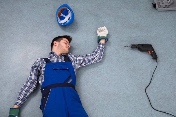 Erhöhter Blick auf bewusstlosen Handwerker, der mit Helm und Bohrer auf dem Boden liegt - Foto, Bild