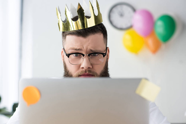 портрет бізнесмена з паперовою короною на голові, дивлячись на екран ноутбука в офісі
 - Фото, зображення