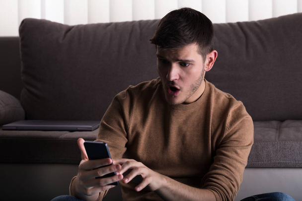 Worried Young Man Looking At Smartphone And Sitting In Front Of Sofa At Home - Foto, imagen