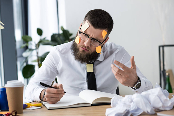 Porträt eines Geschäftsmannes mit klebrigen Notizen auf dem Gesicht, die Notizen in Notizbuch am Arbeitsplatz machen - Foto, Bild