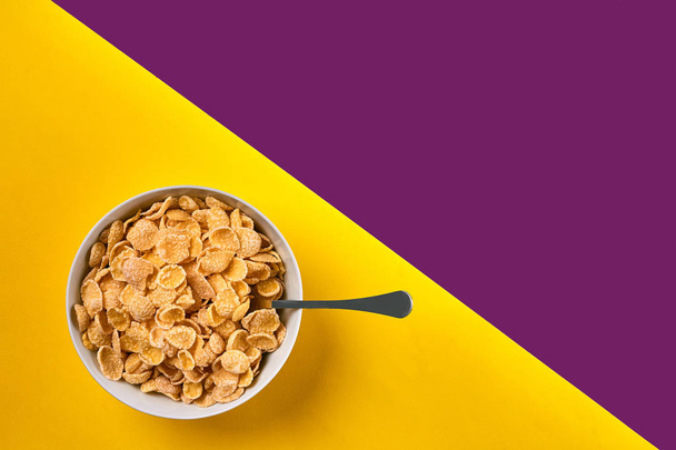Cuenco con hojuelas de maíz y cuchara sobre fondo púrpura y amarillo, vista superior
 - Foto, imagen