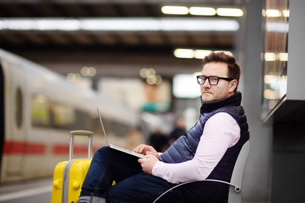 Фрилансер, работающий с ноутбуком на вокзале в ожидании транспорта. Красивый мужчина среднего возраста на платформе железнодорожного вокзала
. - Фото, изображение