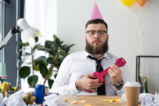 portrait of businessman with party cone on head and toy guitar at workplace in office - Photo, Image