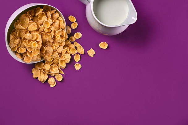 Bowl with corn flakes, jug of milk on purple background, top view - Photo, Image