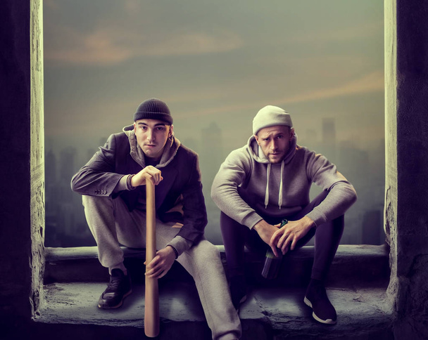Два грабителя сидят на подоконнике. Уличные бандиты с бейсбольной битой и бутылкой алкоголя в ожидании жертвы, концепция преступления
 - Фото, изображение