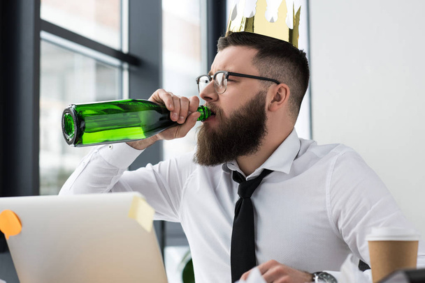 職場のオフィスでシャンパンを飲んで頭に紙の王冠を持ったビジネスマン - 写真・画像