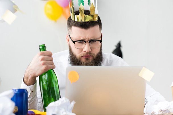 businessman with paper crown on head and bottle of champagne in hand looking at laptop screen at workplace - Zdjęcie, obraz