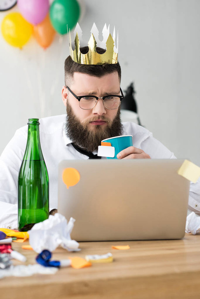 portrait of businessman with paper crown on head looking at laptop screen at workplace in office - Photo, Image