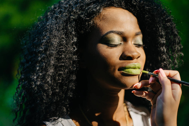 Ruční profesionální vizážistky používat zelený rtěnku pomocí štětce na první pohled okouzlující afro-american dívka s zelené oční stíny na zeleném pozadí. - Fotografie, Obrázek