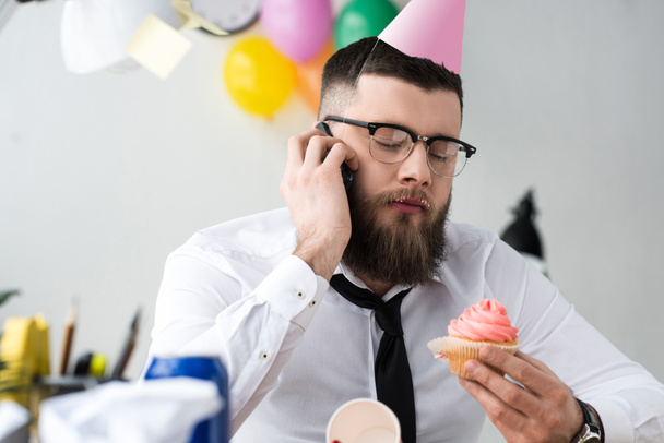 portrait of businessman in party cone with birthday cupcake in hand talking on smartphone - Φωτογραφία, εικόνα