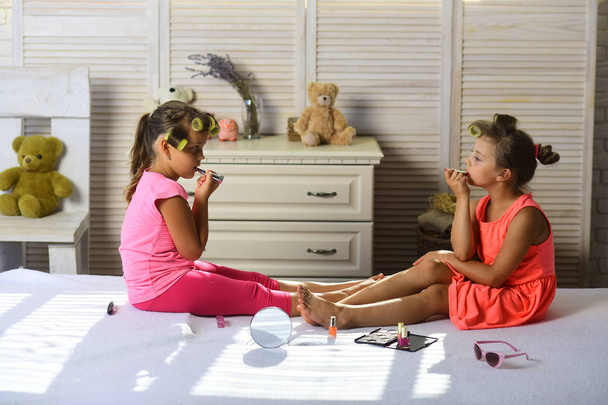Children sit on bed with lipstick, nail polish and mirror. Little girls with serious faces do makeup in room with toys. Girls with curlers play with makeup accessories. Beauty and fashion concept. - Photo, image