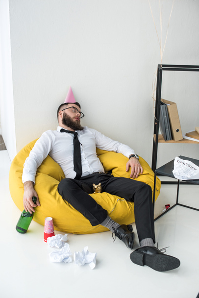 drunk businessman in formal wear with paper cone on head sleeping on bag chair - Foto, imagen