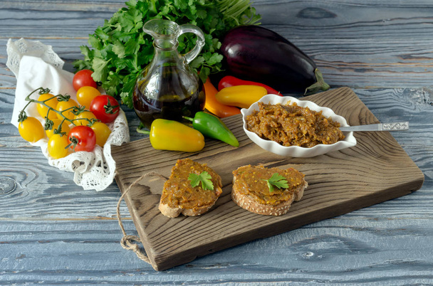 Gesunde Ernährung. gedünsteter Auberginen-Kaviar in einer Schüssel und auf Brotstücken, Kirschtomaten, Petersilie, Pfeffer und Olivenöl in Großaufnahme auf einem Holztisch - Foto, Bild