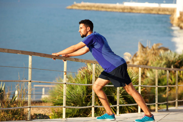 Full length portrait of fit young man stretching muscles by the sea - Photo, Image