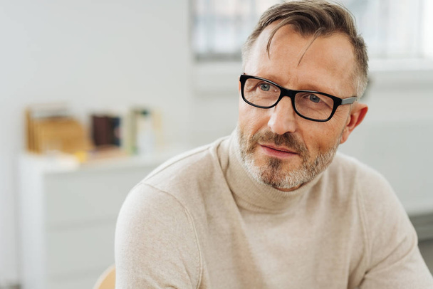 Bearded middle-aged man wearing glasses sitting in an office staring to the side with an attentive expression as though waiting for a response or pondering a problem - 写真・画像