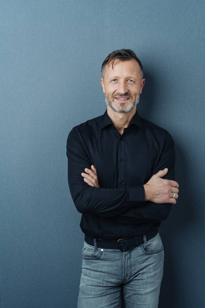 Confident attractive middle-aged man standing with folded arms against a dark studio background smiling at the camera - Valokuva, kuva