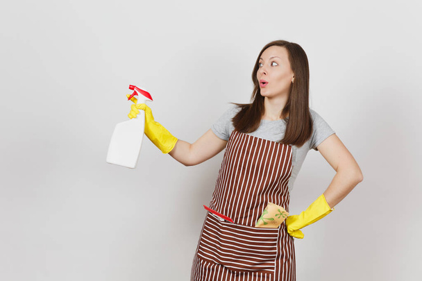 Young smiling housewife in yellow gloves, striped apron, cleaning rag, squeegee in pocket isolated on white background. Housekeeper woman holding spray bottle with cleaner liquid. Bottle copy space. - Valokuva, kuva