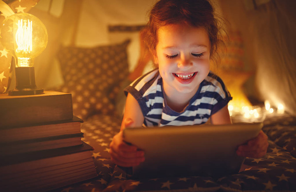 niña feliz con la tableta PC riendo en la oscuridad en la tienda de campaña en casa
 - Foto, imagen