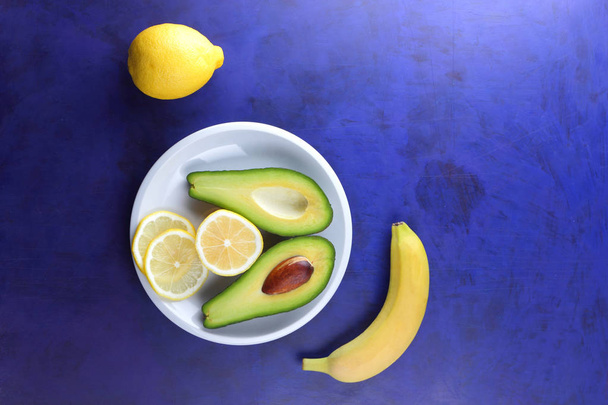 Avocado, banana and lemon on a white plate, tropical fruits on an ultraviolet background, vegetarian breakfast, Asian cuisine, avocado pop art, citrus and banana with avocado - Photo, image