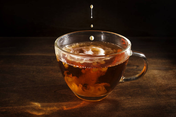 black tea freshly brewed in a glass cup and some drops of milk or cream against a dark rustic wooden background, hot drink against cold and flu, copy space - Foto, Imagem