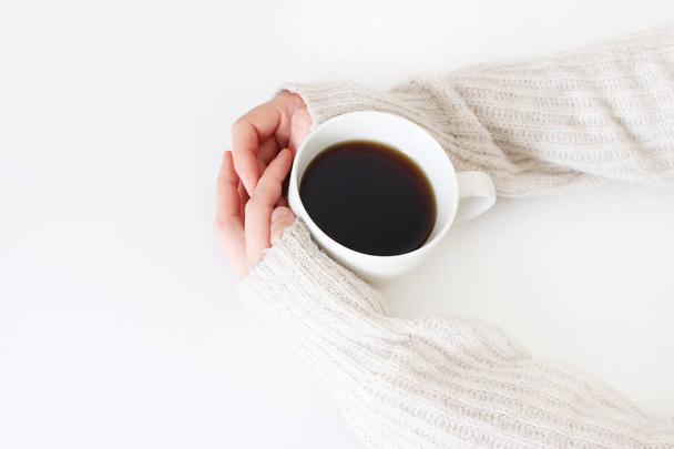 Womans hands in sweater holding cup of coffee on white table. Top view, styled stock image. Break, breakfast concept. - Фото, изображение