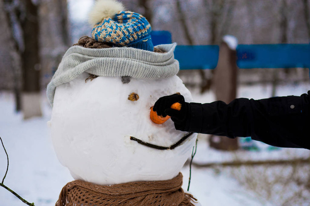  Die Hand des Mannes hält eine große Karotte, die Nase eines echten großen Schneemanns im Winterpark, Winterzeit - Foto, Bild