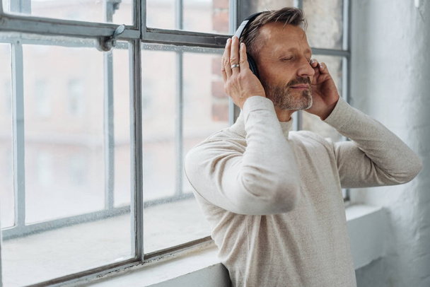 Attractive middle-aged man listening to music on stereo headphones as he leans back against a bright window - Foto, afbeelding