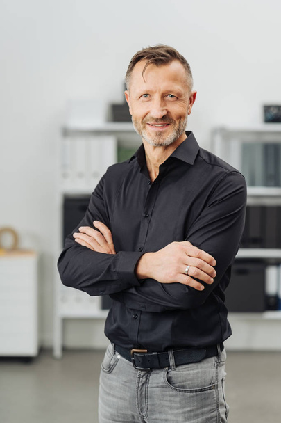 Friendly self-assured middle-aged man standing with crossed arms in a large office smiling at the camera - Fotografie, Obrázek