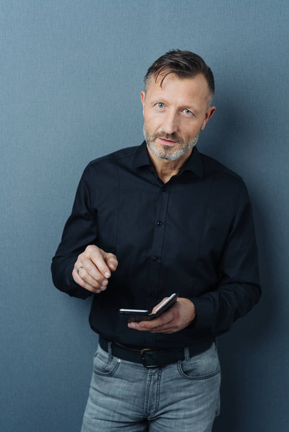 Serious thoughtful businessman holding a smartphone looking up at the camera with an attentive look against a dark grey studio background - Photo, Image