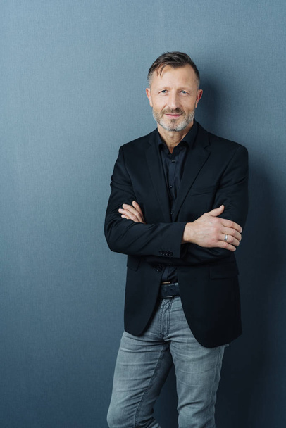 Confident middle-aged man in jeans and jacket standing with folded arms against a dark studio background with copy space - Foto, Bild