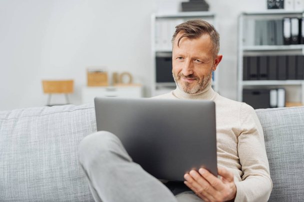 Attractive middle-aged man sitting using a laptop with a pleased smile as he relaxes on a comfortable sofa at home - Foto, immagini