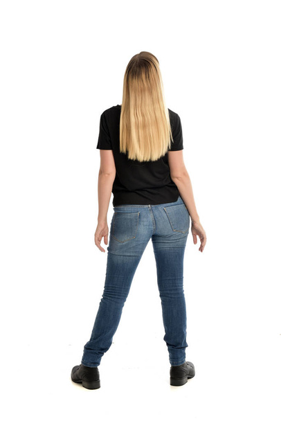 full length portrait of blonde girl wearing simple black shirt and jeans. standing pose facing away from camera, isolated on white background. - Photo, Image