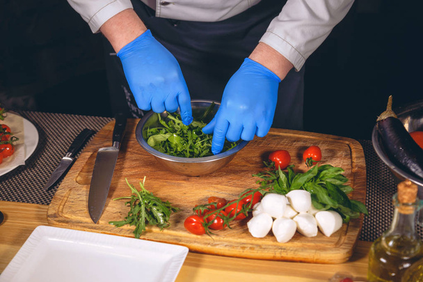 Les mains du chef avec des gants cuits. Chef prépare un plat gastronomique - mozzarella au basilic, tomates cerises et roquette
.  - Photo, image