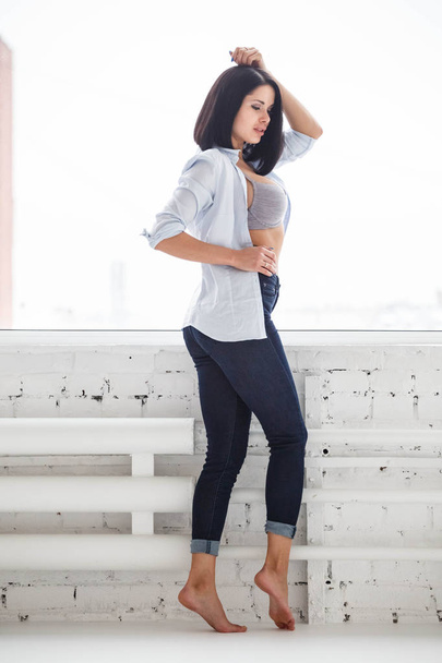 Studio shot of beautiful brunette woman wearing dark jeans and white shirt standing near window at apartment interior - Фото, зображення