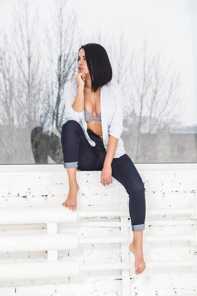 Studio shot of beautiful brunette woman wearing dark jeans and white shirt sitting on windowsill near window at apartment interior - Φωτογραφία, εικόνα